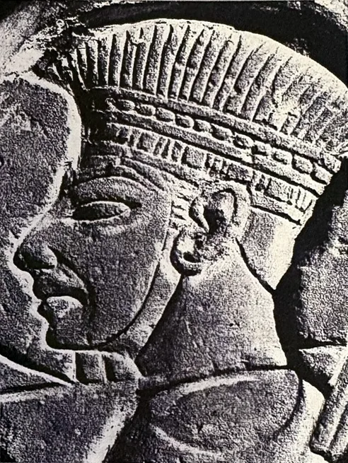 Schwarzweißfoto eines Steinreliefs: Kopf eines Mannes im Profil nach links mit hoher, federartig gerippter Kopfbedeckung und Stirnband.