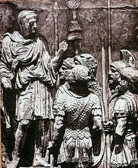 Schwarzweißfoto eines antiken Steinreliefs: Links steht ein Mann in Tunika und Mantel und hebt die rechte Hand; vor ihm stehen dicht gedrängt mehrere Soldaten mit Schuppenpanzern und Helmen, im Hintergrund Feldzeichen und Lanzen.