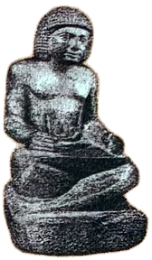 Foto einer kleinen dunklen Steinfigur: Ein Mann mit Kopftuch sitzt mit untergeschlagenen Beinen auf einem Sockel, die Hände auf dem Schoß.