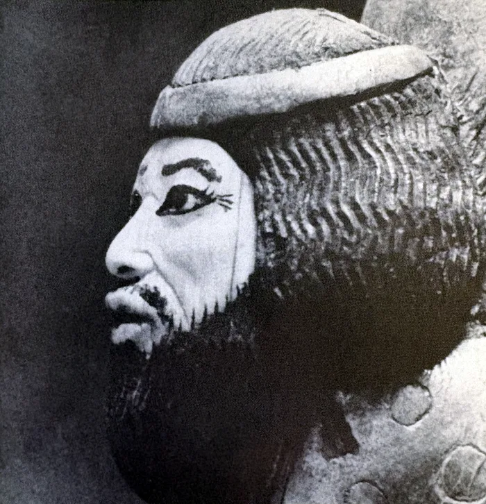 Schwarzweißfoto eines Reliefs oder einer Skulptur: Kopf eines bärtigen Mannes im Profil nach links mit Stirnband, schulterlangem gewelltem Haar und Vollbart; Augen und Brauen sind dunkel umrandet.