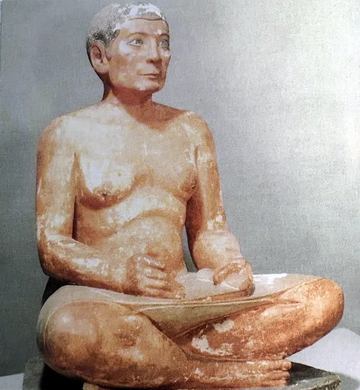 Farbfoto einer bemalten Sitzfigur vor grauem Hintergrund: Ein Mann mit kurzem schwarzem Haar und dunkel umrandeten Augen sitzt mit gekreuzten Beinen, auf dem Schoß eine aufgerollte Schriftrolle, die Hände in Schreibhaltung; er trägt einen weißen Schurz.