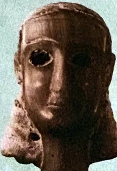 Farbfoto eines Frauenkopfes aus rötlichem Stein oder Bronze, leicht nach links gewandt, mit glatt anliegendem Haar und tief ausgehöhlten Augen; Oberfläche an Wange und Hals beschädigt.