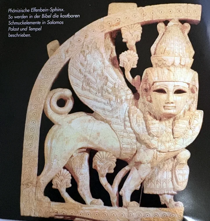 Farbfoto einer durchbrochen geschnitzten Elfenbeintafel vor schwarzem Grund: Eine geflügelte Sphinx mit menschlichem Gesicht, hoher Krone und Löwenkörper schreitet nach rechts zwischen stilisierten Pflanzen, eingefasst von einem Bogen mit Kreisornamenten. Oben links ist die Bildunterschrift in das Foto gedruckt.